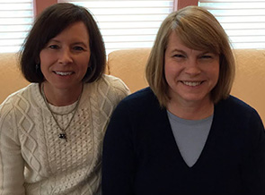 Two women smiling indoors, sitting closely together.